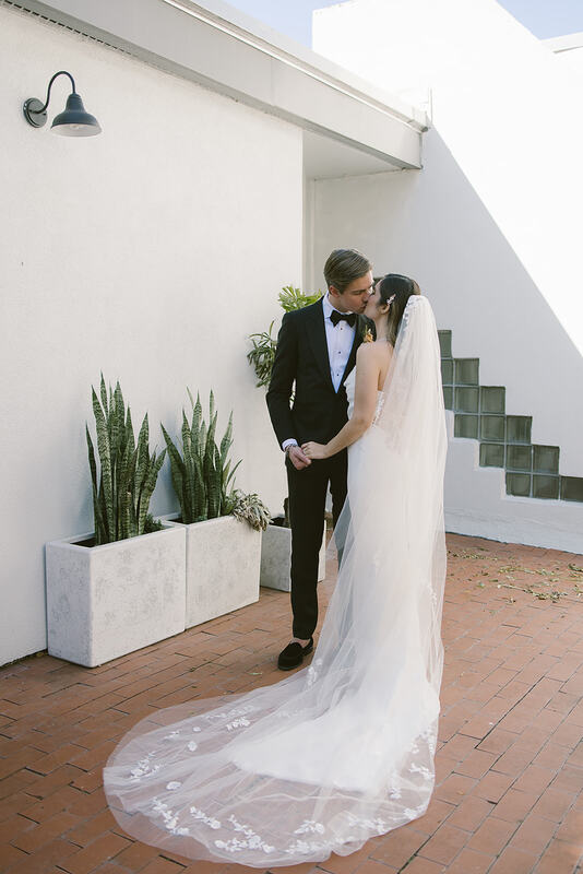 Bride wearing a sleek, elegant wedding gown and a detailed veil, kissing her groom who is wearing a classic black tux, crips white shirt, and black bow-tie. They are standing in a courtyard surrounded by white walls, terracotta cobblestone, and snake plants in a grey, marble, pots. 